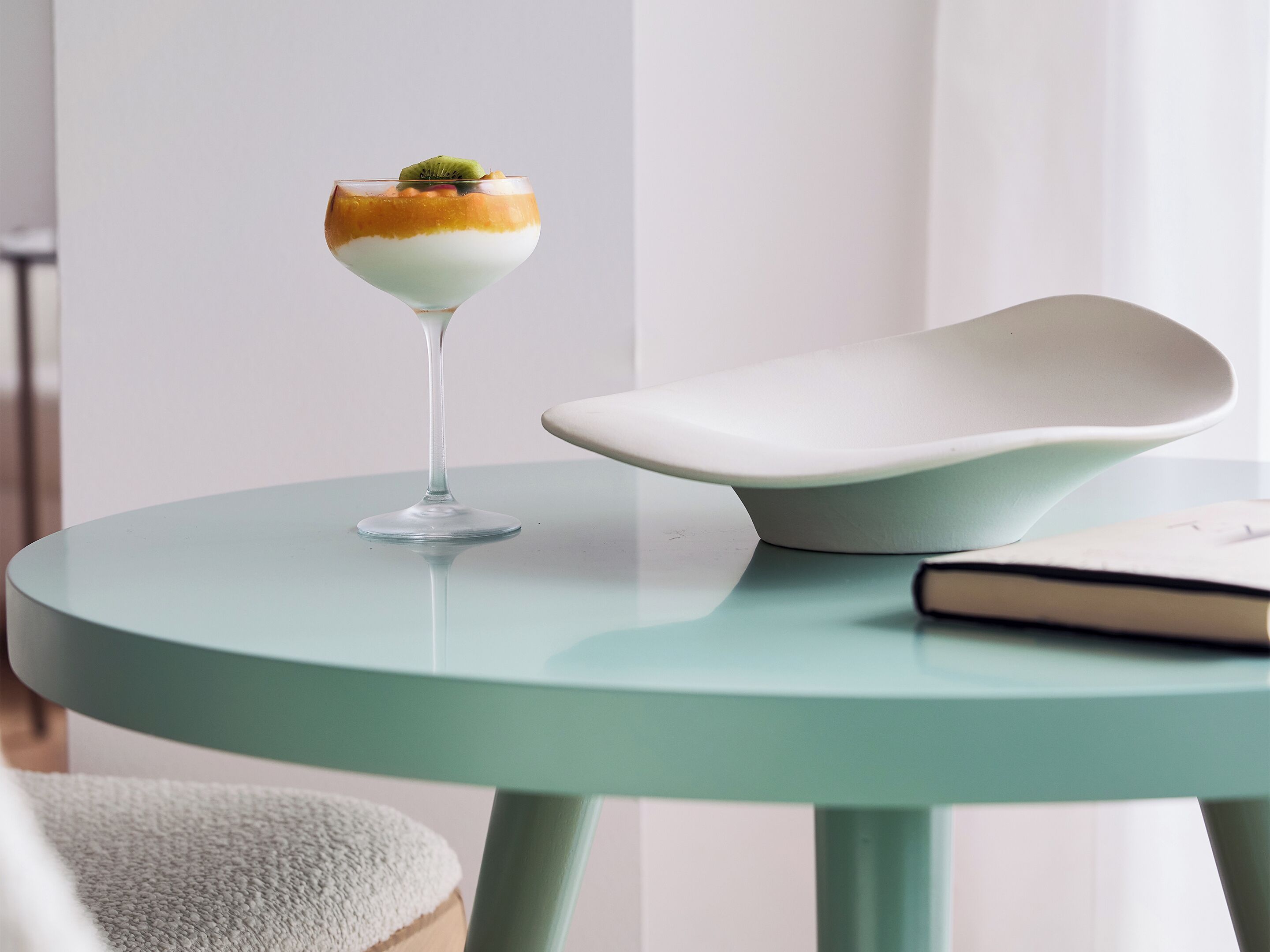 A dessert in a glass on a teal table with a modern chair and a book beside it.