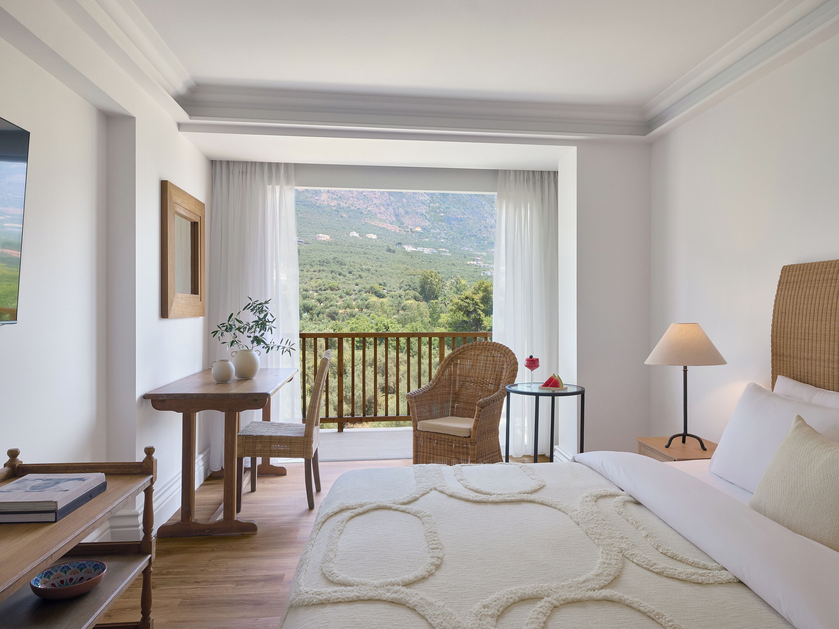 A hotel room with a bed, a table with a bowl, a chair, a lamp, and a view of a mountainous landscape.