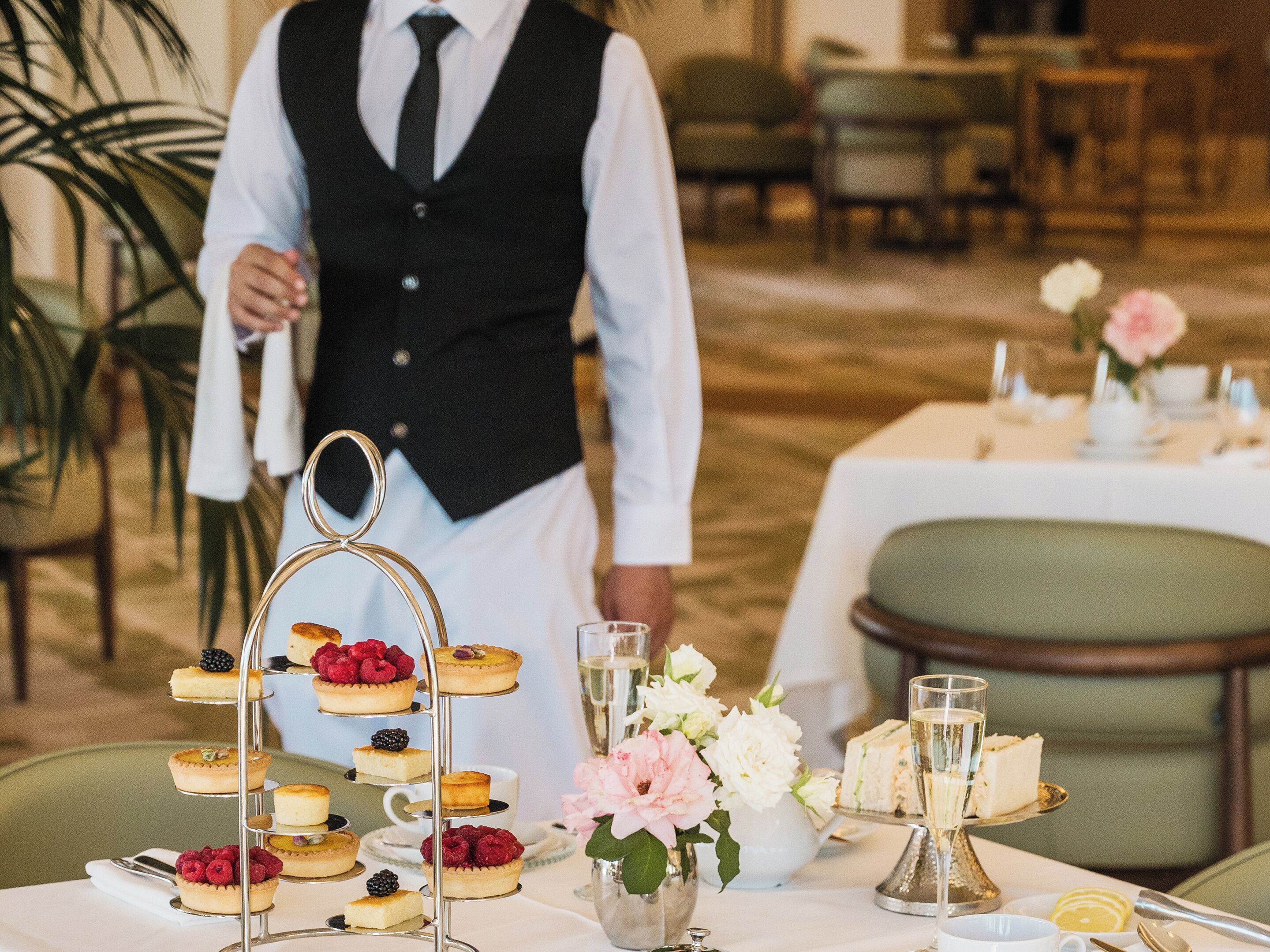 A formal dining setting with a tiered dessert stand, a waiter, and a table set with flowers and glasses.