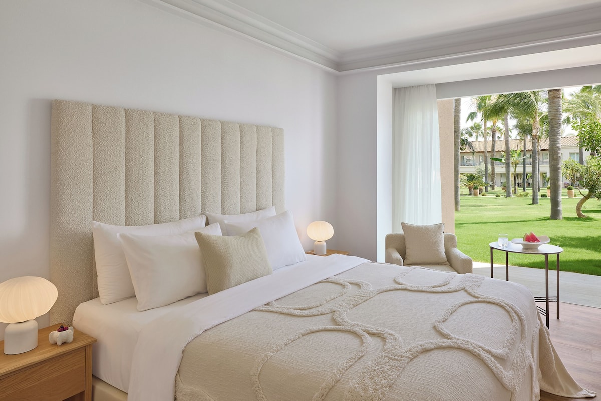 Une chambre d’hôtel avec un grand lit, une table de chevet avec une lampe, une chaise et une vue sur une cour intérieure agrémentée de palmiers.