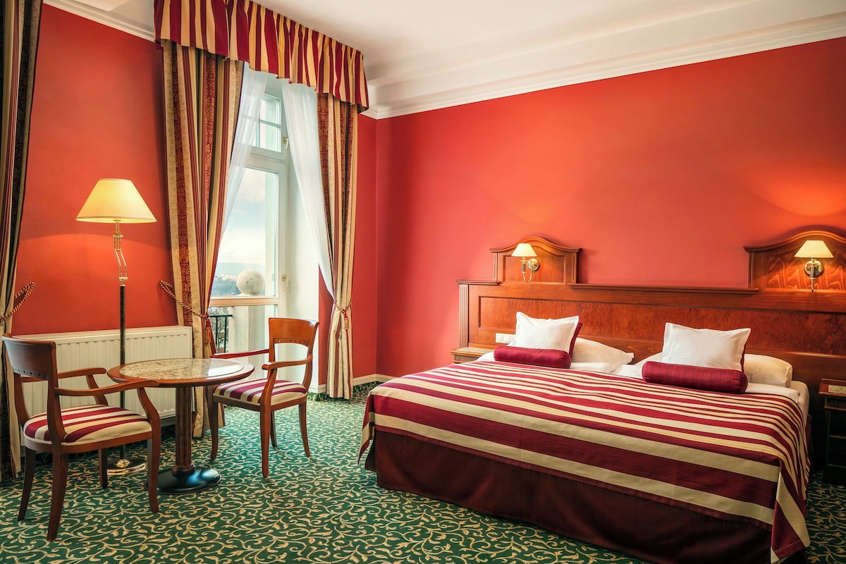 Una habitación de hotel con una pared roja, una cama con ropa de cama a rayas, un cabecero de madera, dos lámparas de noche, una mesa redonda con sillas y una alfombra estampada.