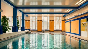 Una piscina interior con agua cristalina, paredes azules y anaranjadas, y ventanas amplias.