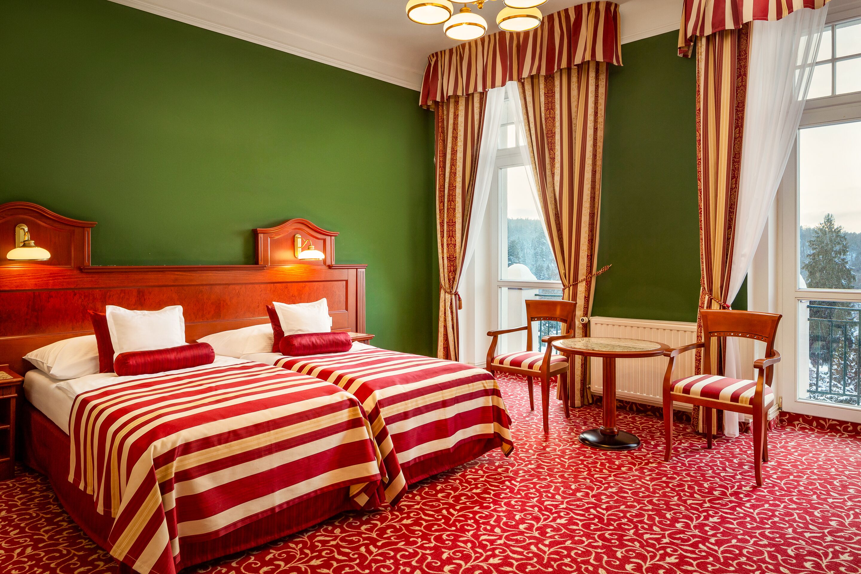 Ein Hotelzimmer mit zwei Betten, einem Holzkopfteil, einem Nachttisch mit Lampe, einem Sessel, einem kleinen Tisch und Blick auf Bäume durch das Fenster.