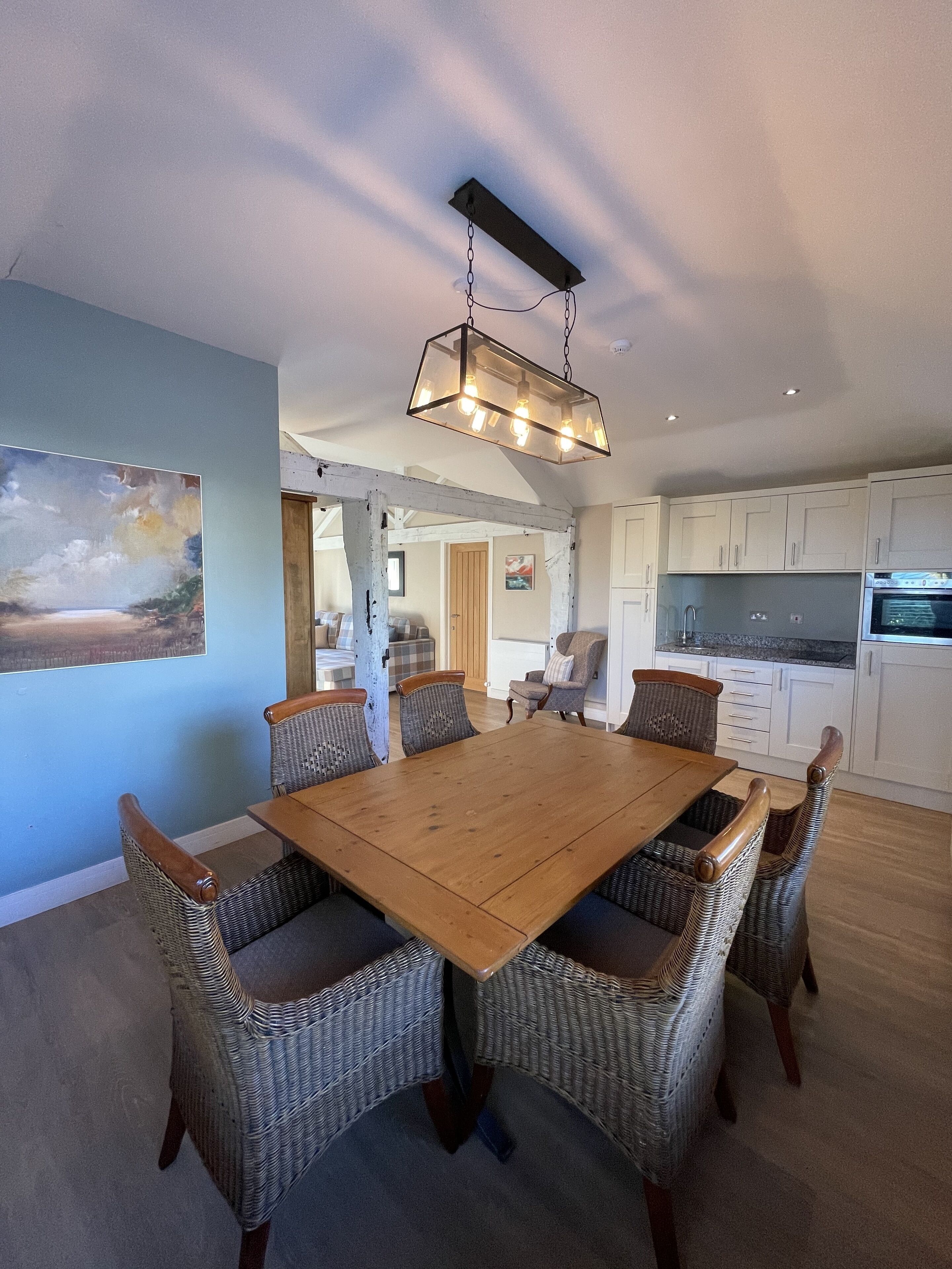 A dining area with a wooden table and wicker chairs, a kitchen with white cabinets, and a painting on the wall.