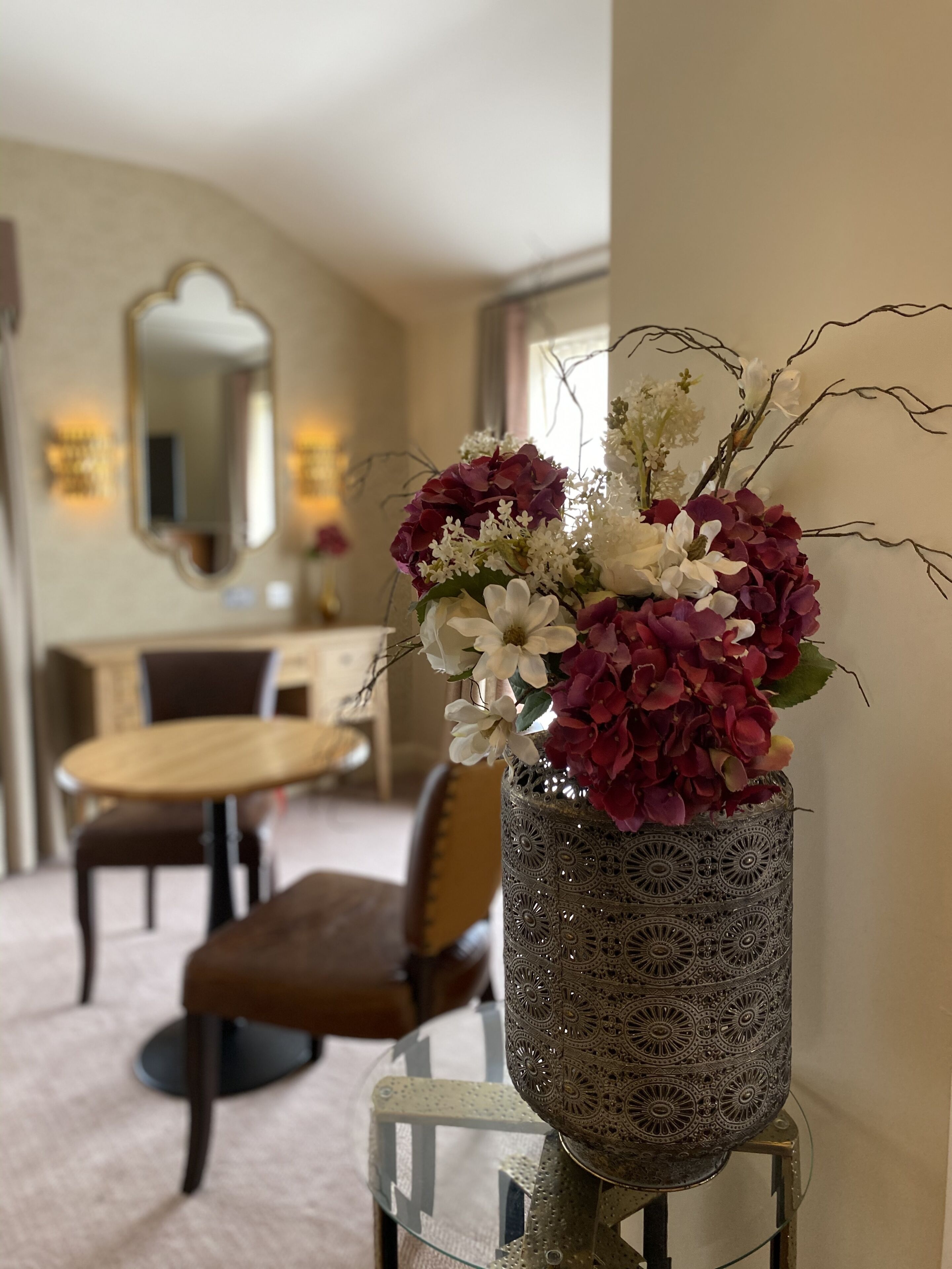 A dining area with a table and chairs, a decorative vase with flowers, and a mirror on the wall.