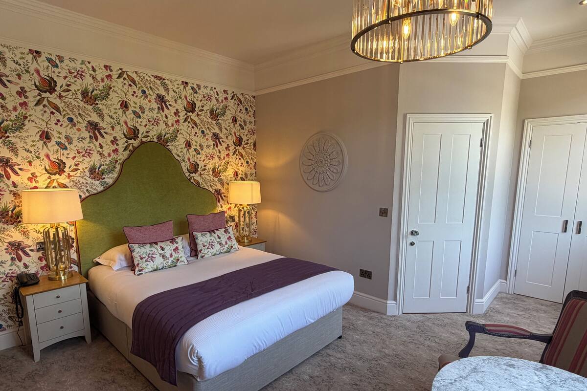 Een slaapkamer met een bed, twee nachtkastjes, een lamp, een stoel, een ronde tafel en een deur.