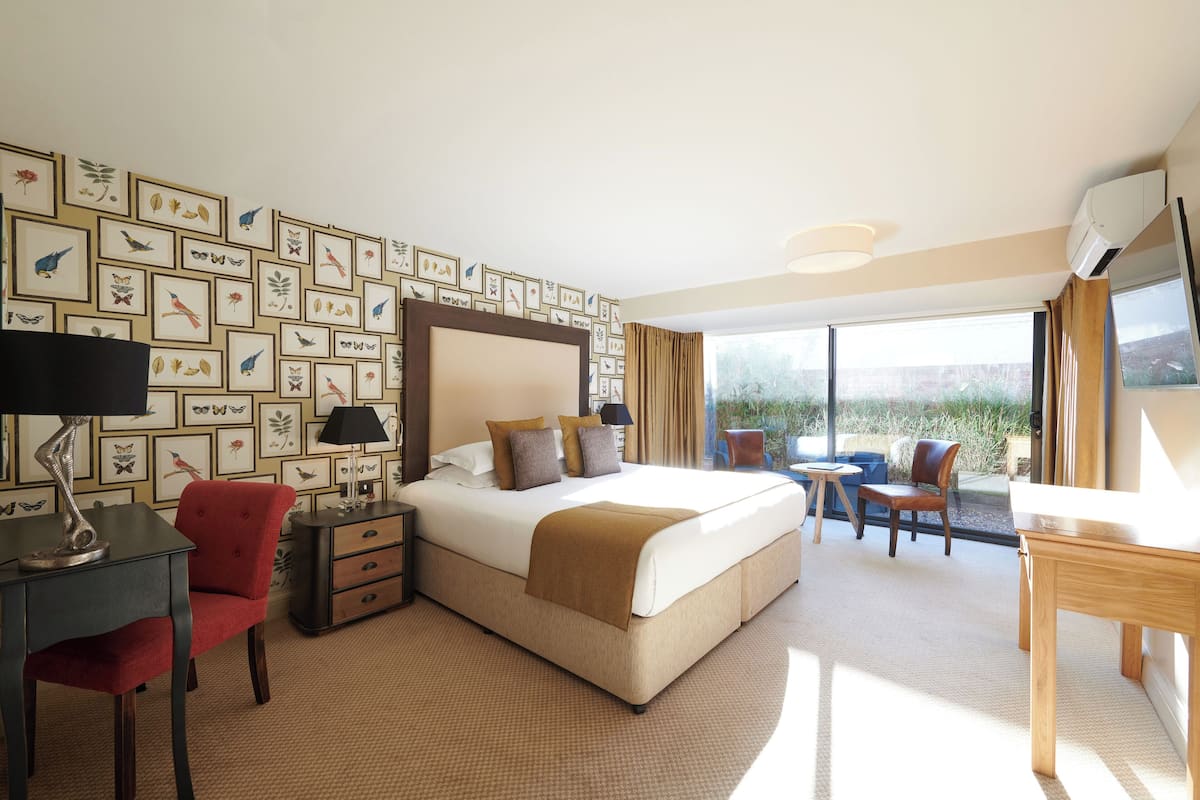 Een hotelkamer met een groot bed, een bureau met lamp, een stoel, een klein tafeltje en uitzicht op het landschap door een groot raam.