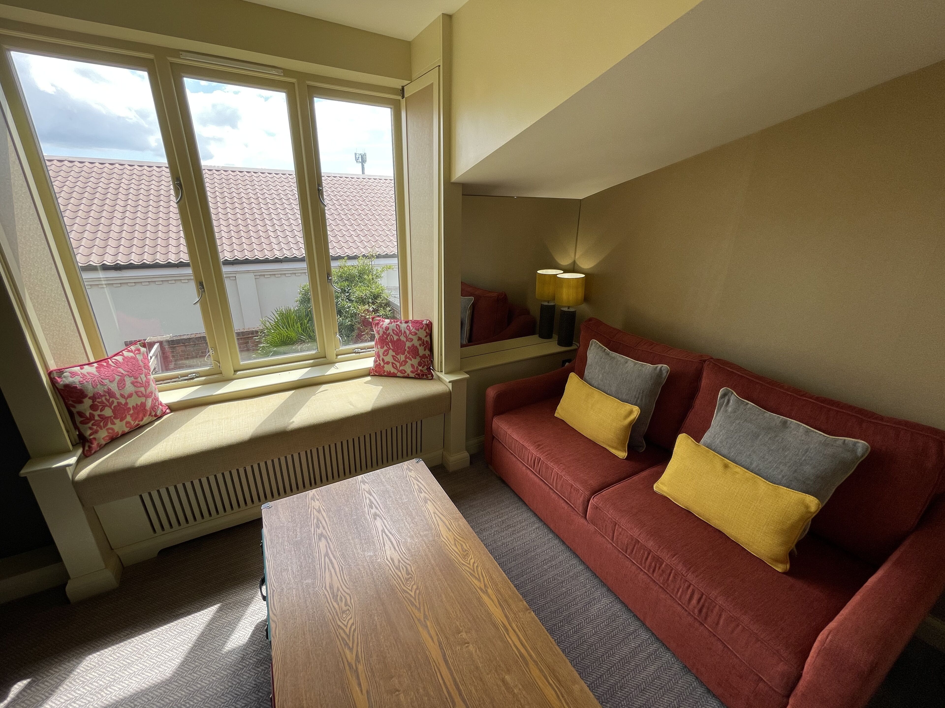A cozy living room with a red sofa, a wooden coffee table, and a window with a view of a red-tiled roof.