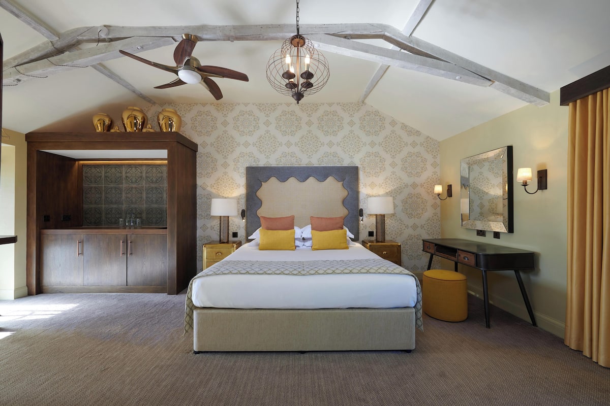 Een ruime slaapkamer met een groot bed, een houten garderobe, een bureau met stoel en een plafondventilator.