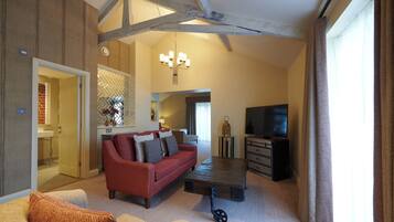A spacious living room with a red sofa, a wooden coffee table, and a television. There is a kitchen area visible through an open door.