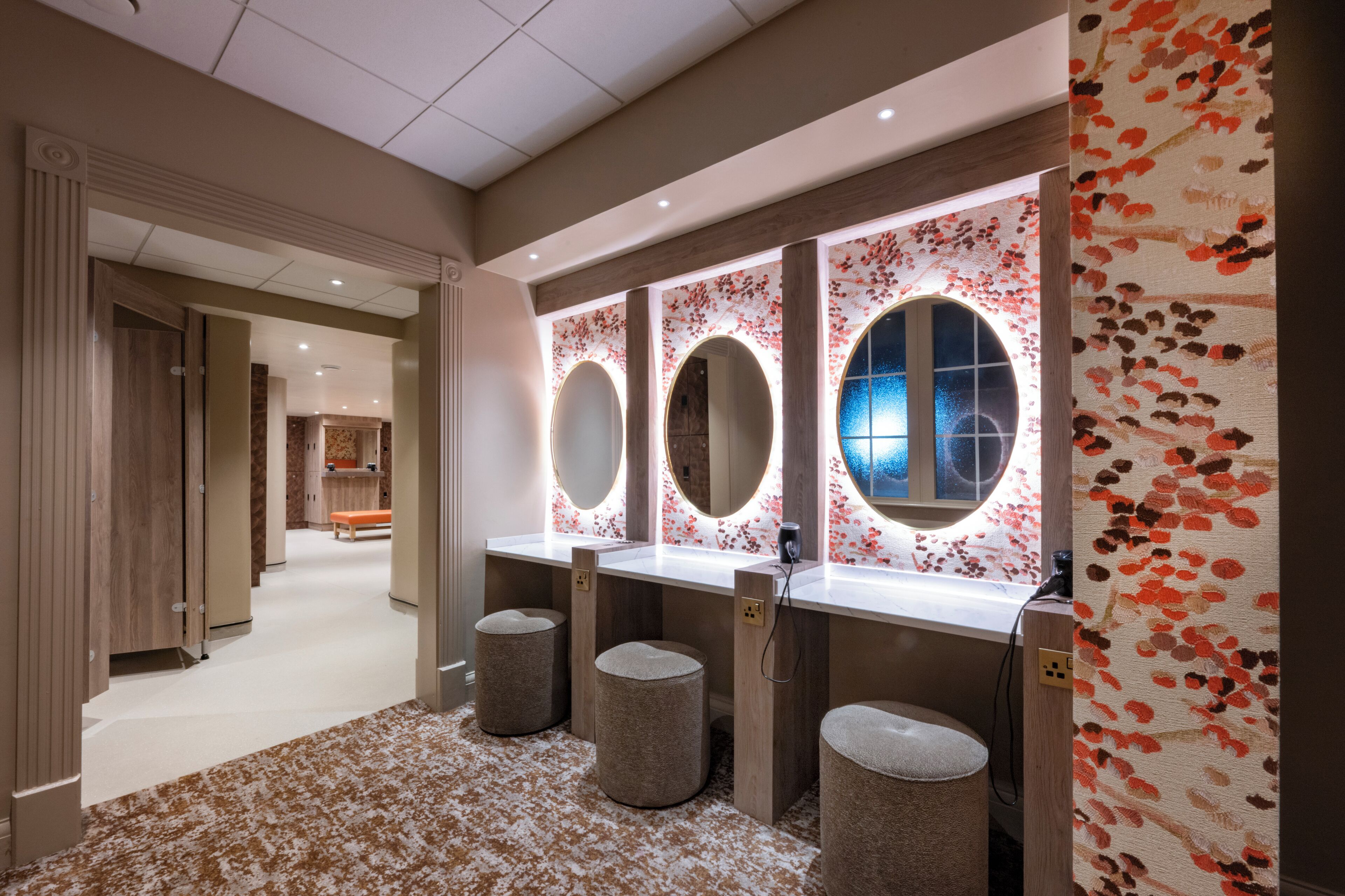 A modern bathroom with three sinks, each with a round mirror and underlighting. There are three stools and a bench in the space.