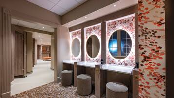A modern bathroom with three sinks, each with a round mirror and underlighting. There are three stools and a bench in the space.