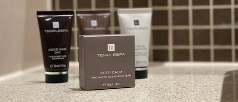 A bathroom with a tiled wall and a countertop. On the countertop, there are three Temple Spa products: a black tube labeled "GOOD HAIR DAY SHAMPOO," a brown tube labeled "TEMPLESPA," and a white tube labeled "TEMPLESPA." In front of these products is a Temple Spa box labeled "KEEP CALM AROMATIC CLEANSING BAR."