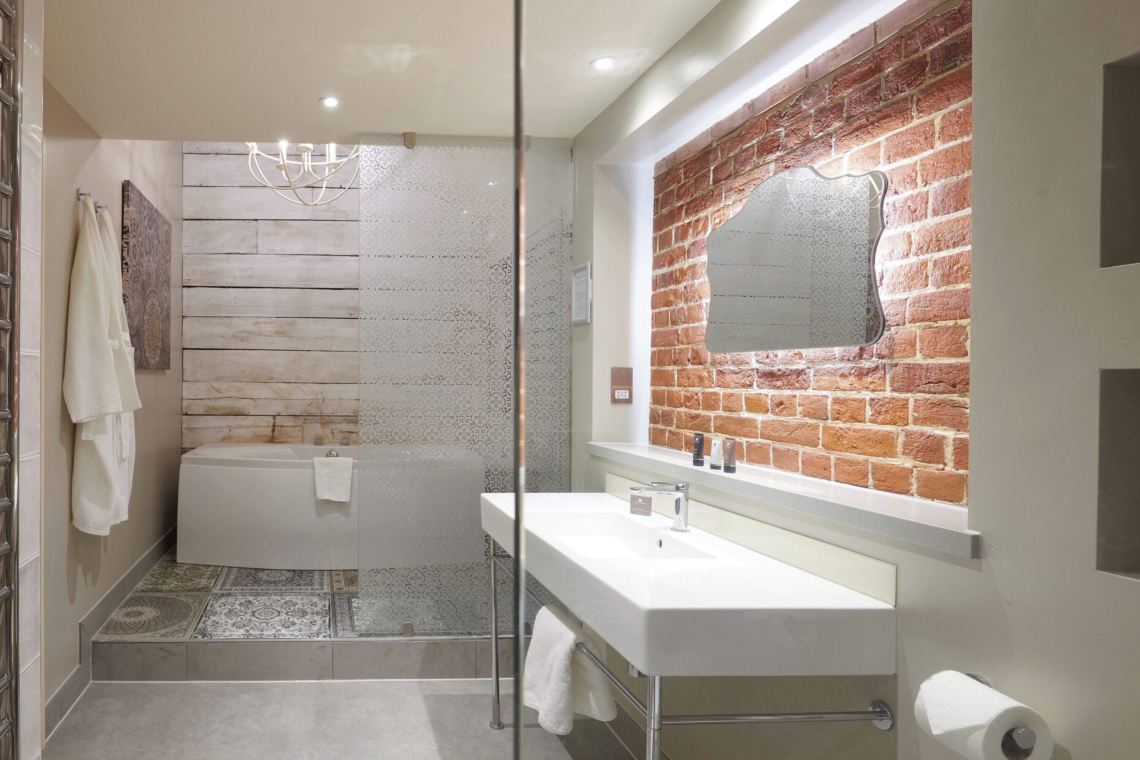 A modern bathroom with a bathtub, a glass-enclosed shower, a white sink, and a mirror with a brick wall backdrop.