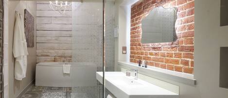 A modern bathroom with a bathtub, a glass-enclosed shower, a white sink, and a mirror with a brick wall backdrop.