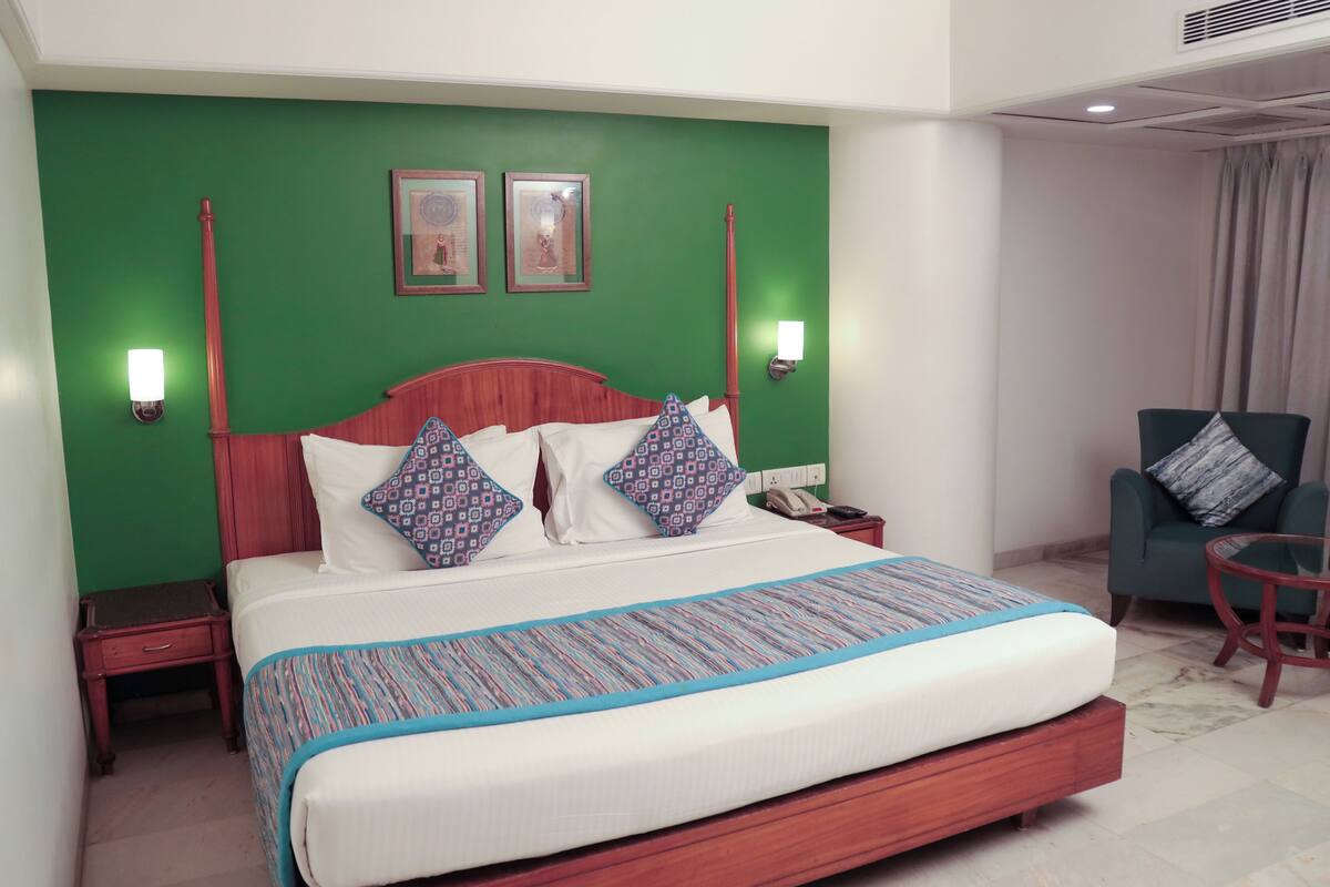Habitación de hotel con una cama grande, paredes verdes, dos cuadros enmarcados, una mesita de noche con un teléfono y un sillón verde.