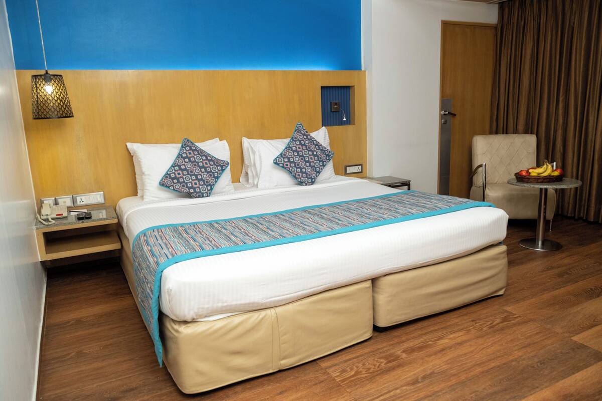 Una habitación de hotel con una cama grande, una silla, una mesita con fruta y una lámpara de pared.