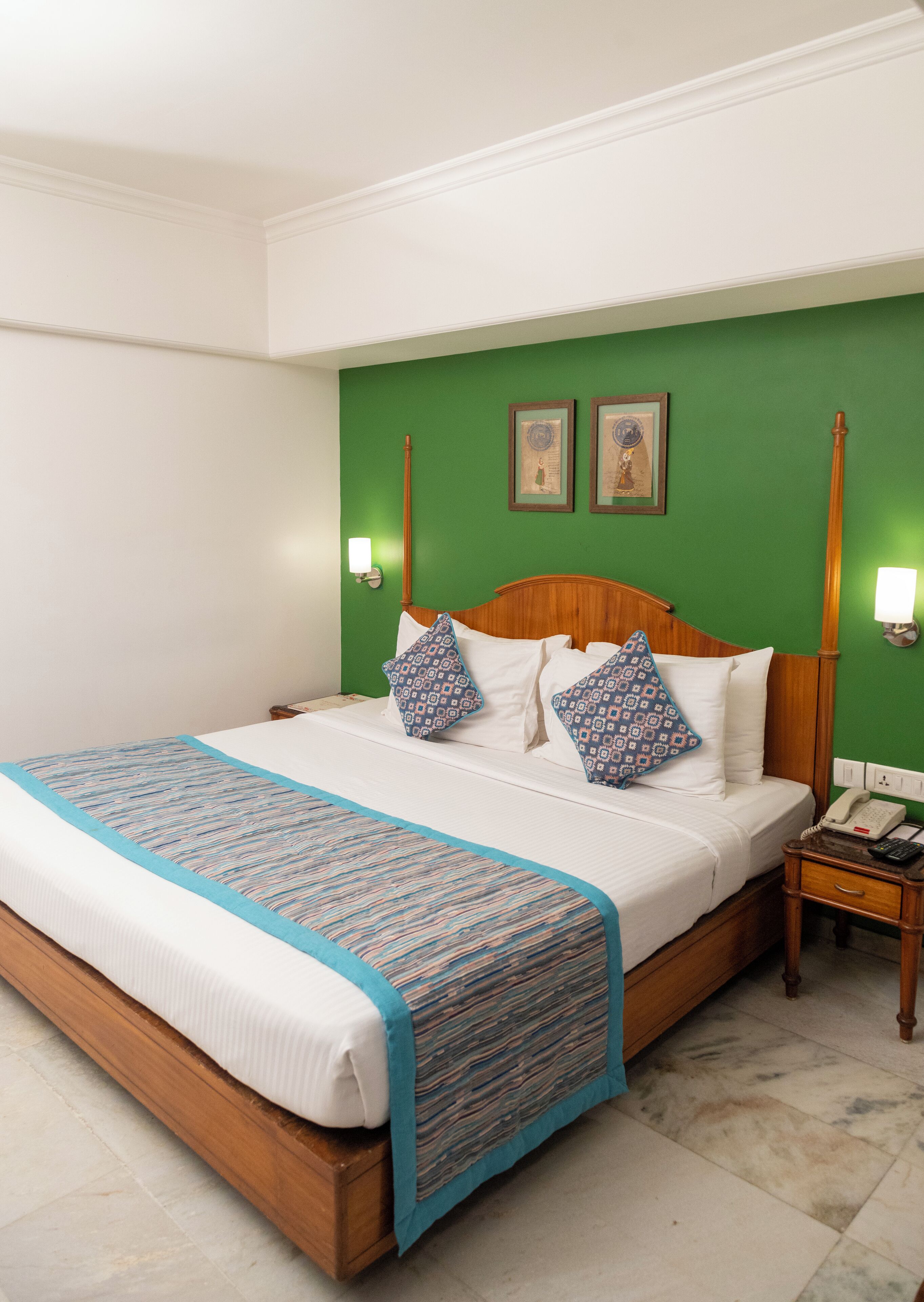 Ein Hotelzimmer mit einem großen Bett, grünen Wänden, zwei gerahmten Bildern, einem Nachttisch mit Telefon und einem bei dem Bett stehenden Tisch.