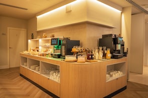 Un sector de desayuno autoservicio con diversas opciones de alimentos, una cafetera y un menú digital.
