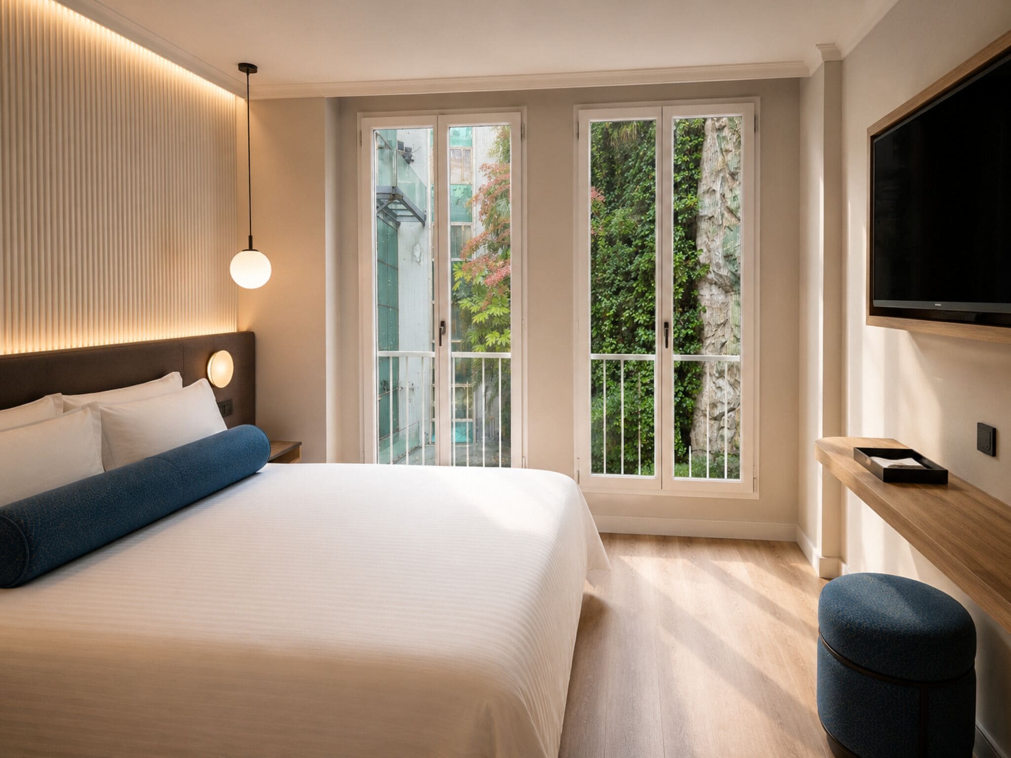 A hotel room with a bed, a TV, a desk, and a window with a view of greenery.