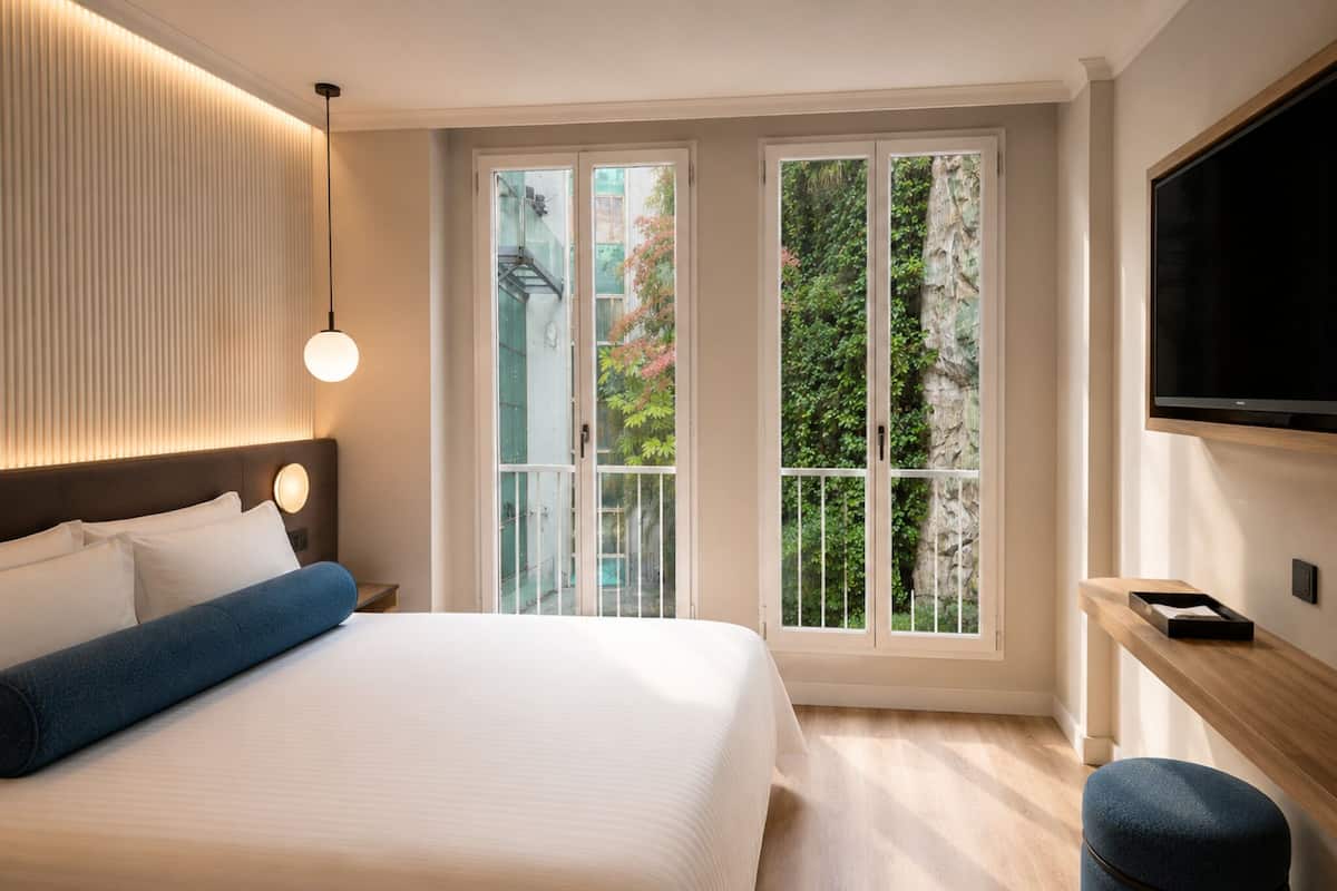 Ein Hotelzimmer mit Bett, Fernseher, Schreibtisch und Fenster mit Blick auf Grünflächen.