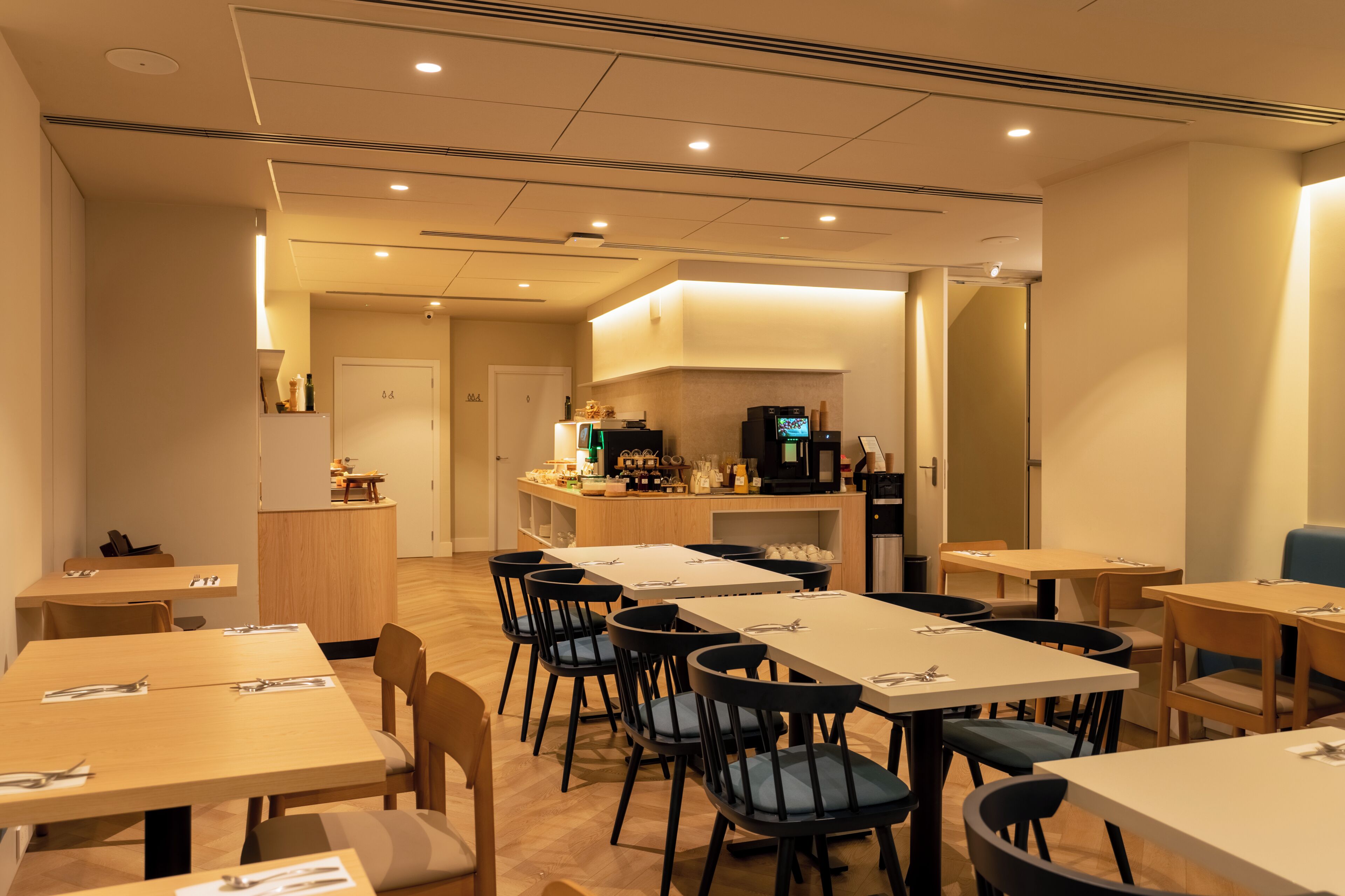 Un comedor de hotel con varias mesas y sillas, una cocina al fondo y un diseño interior moderno.