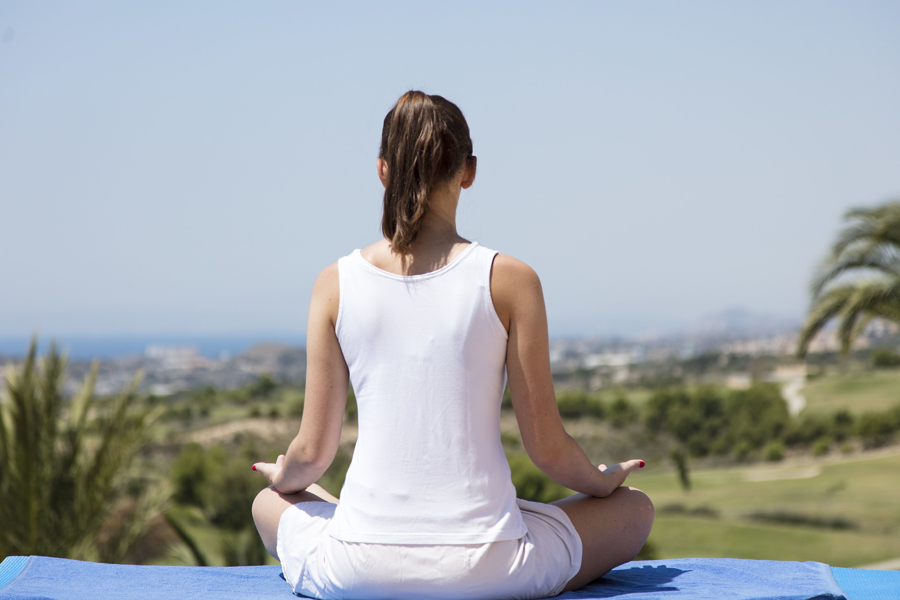 Una persona sentada en una esterilla de yoga al aire libre, con vistas a un paisaje urbano y zonas verdes al fondo.