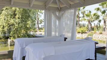Dos camas blancas con ropa de cama blanca, una terraza de madera y vistas a palmeras y una masa de agua.