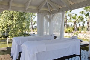 Two white beds with white linens, a wooden deck, and a view of palm trees and a body of water.