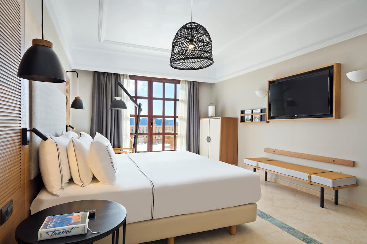 A hotel room with a bed, bedside table, lamp, TV, bench, and a view of the outdoors.