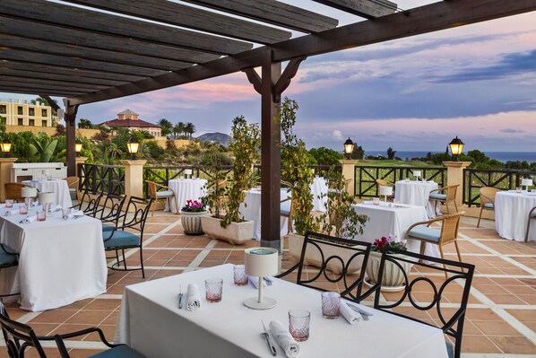 Un área de comedor al aire libre con mesas y sillas, con vistas a un paisaje montañoso y una masa de agua.