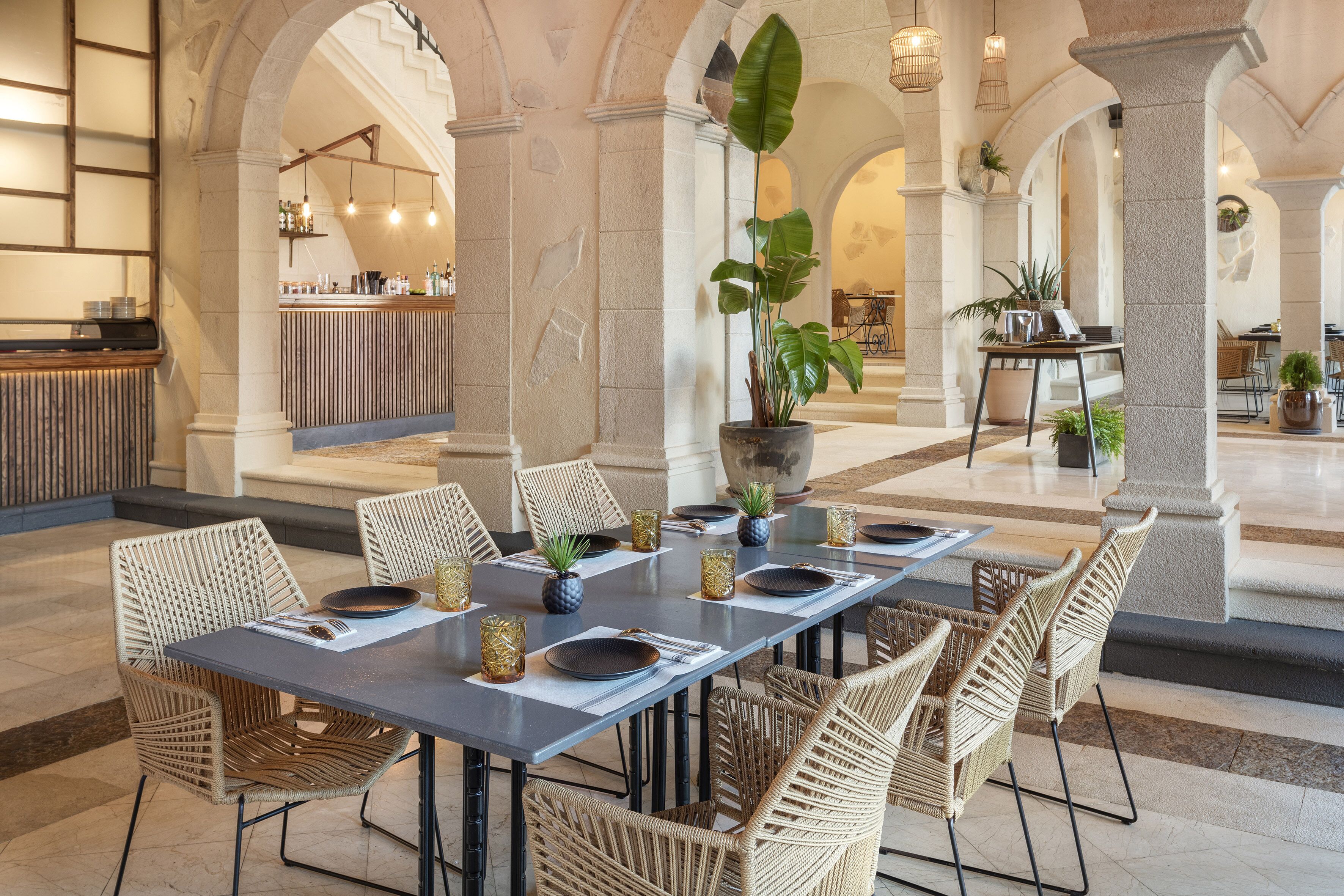 Un comedor amplio con una mesa larga dispuesta para seis personas, rodeada de sillas de mimbre. Se observan plantas en macetas y una zona de bar al fondo.