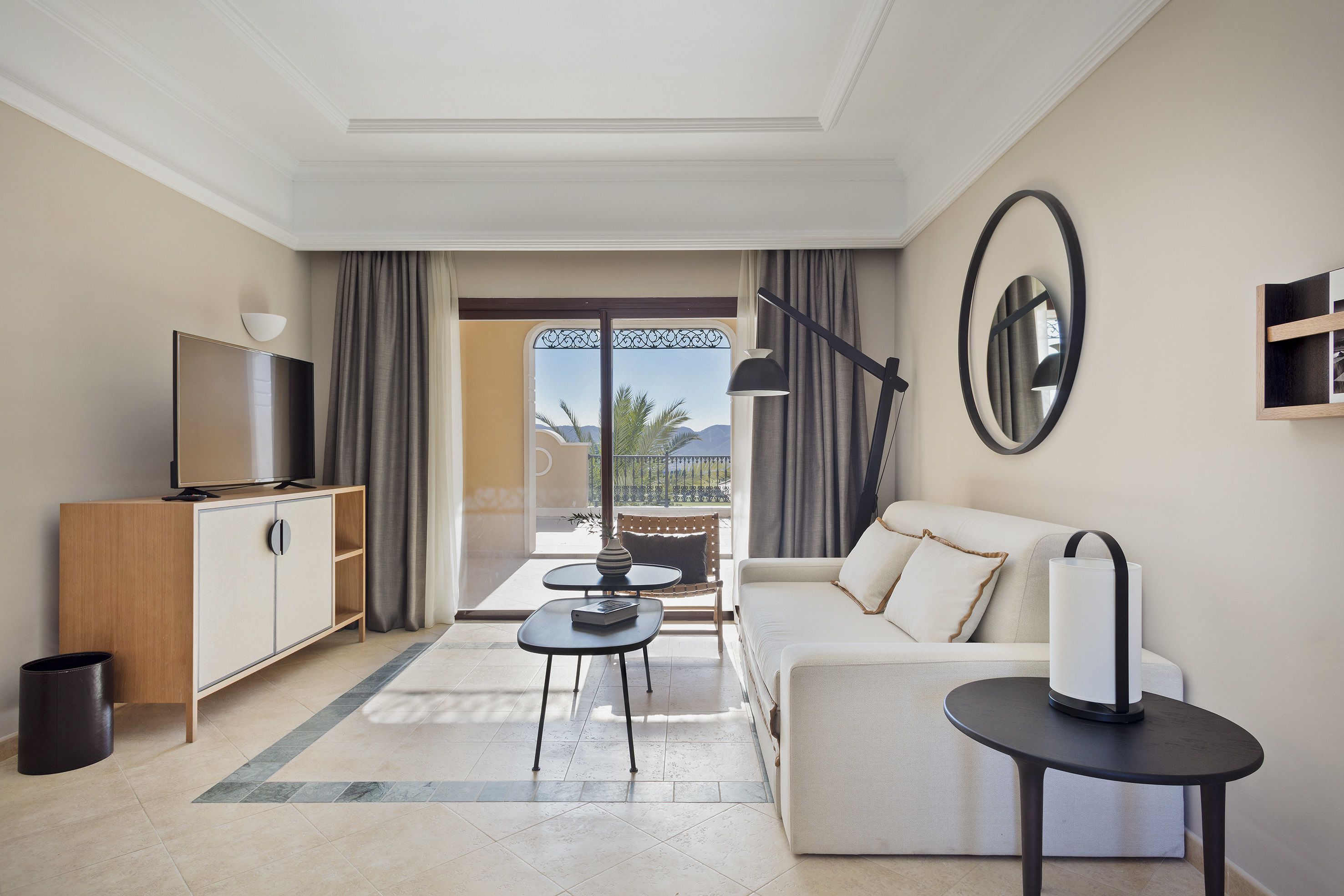 A modern hotel room with a sofa, a TV, a small table, and a balcony with a view.