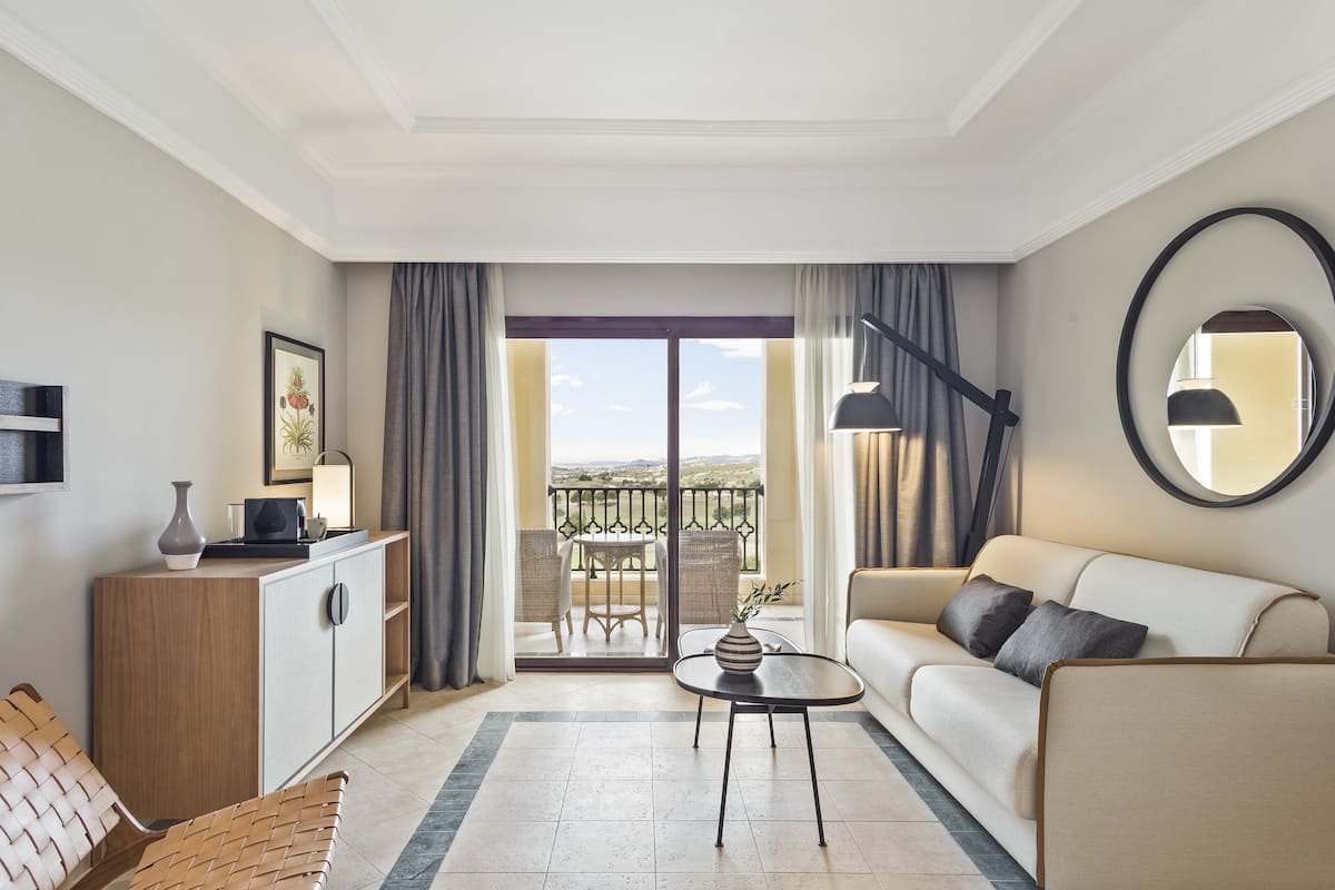 A modern hotel room with a sofa, a small table with a vase, a cabinet, a round mirror, and a balcony with a view.