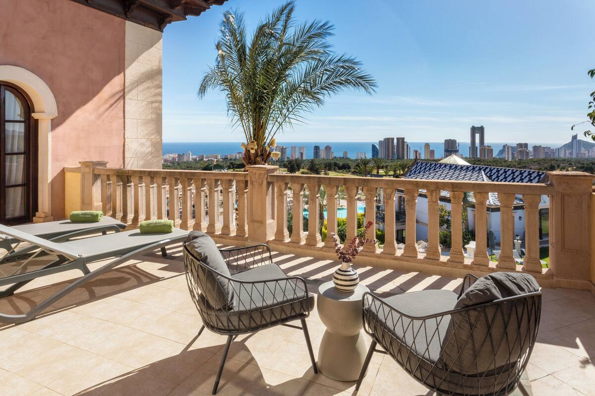 A balcony with a view of the city skyline, a palm tree, and outdoor seating.