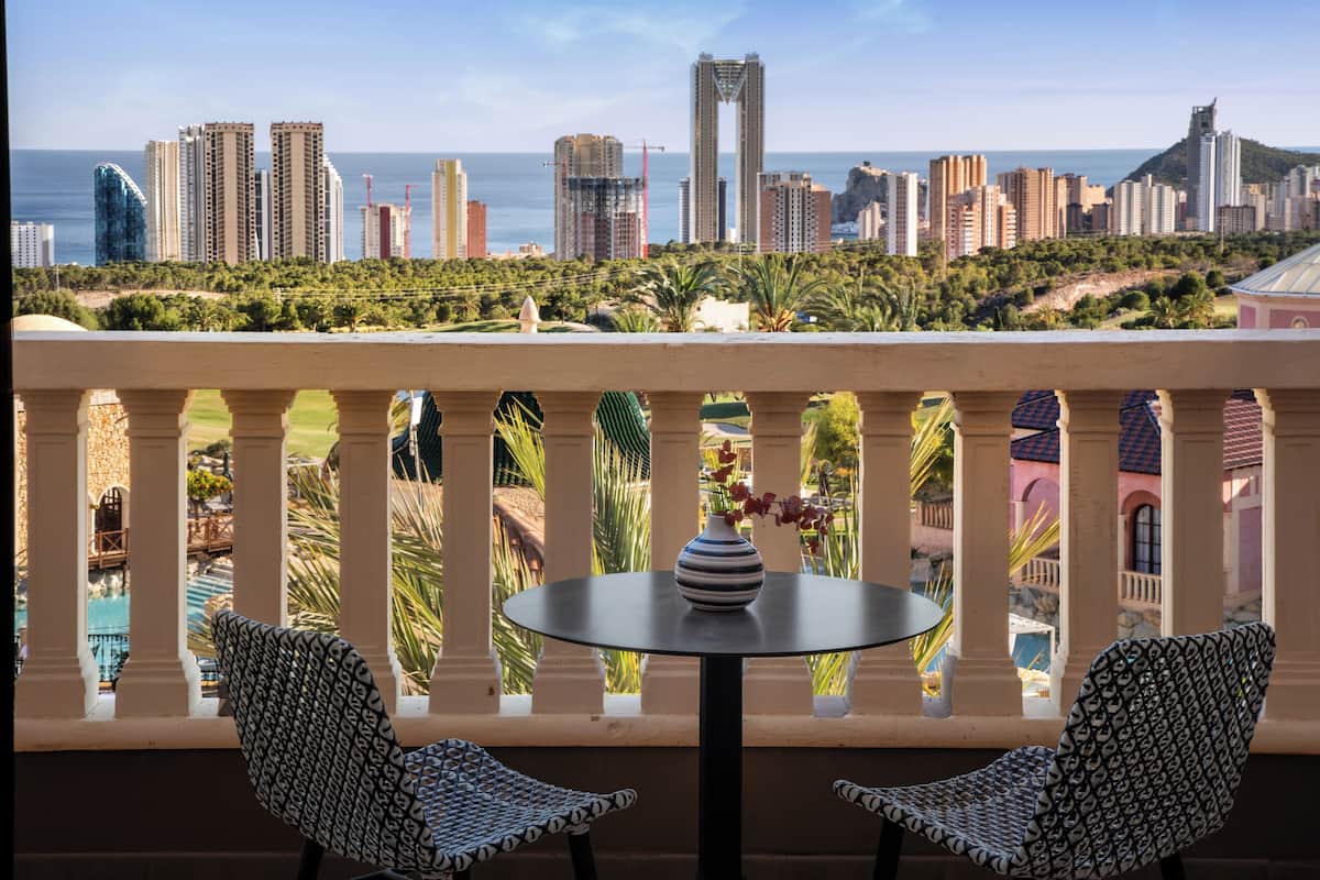 A balcony with two patterned chairs, a small table with a vase, and a view of a city skyline with high-rise buildings and a beach.