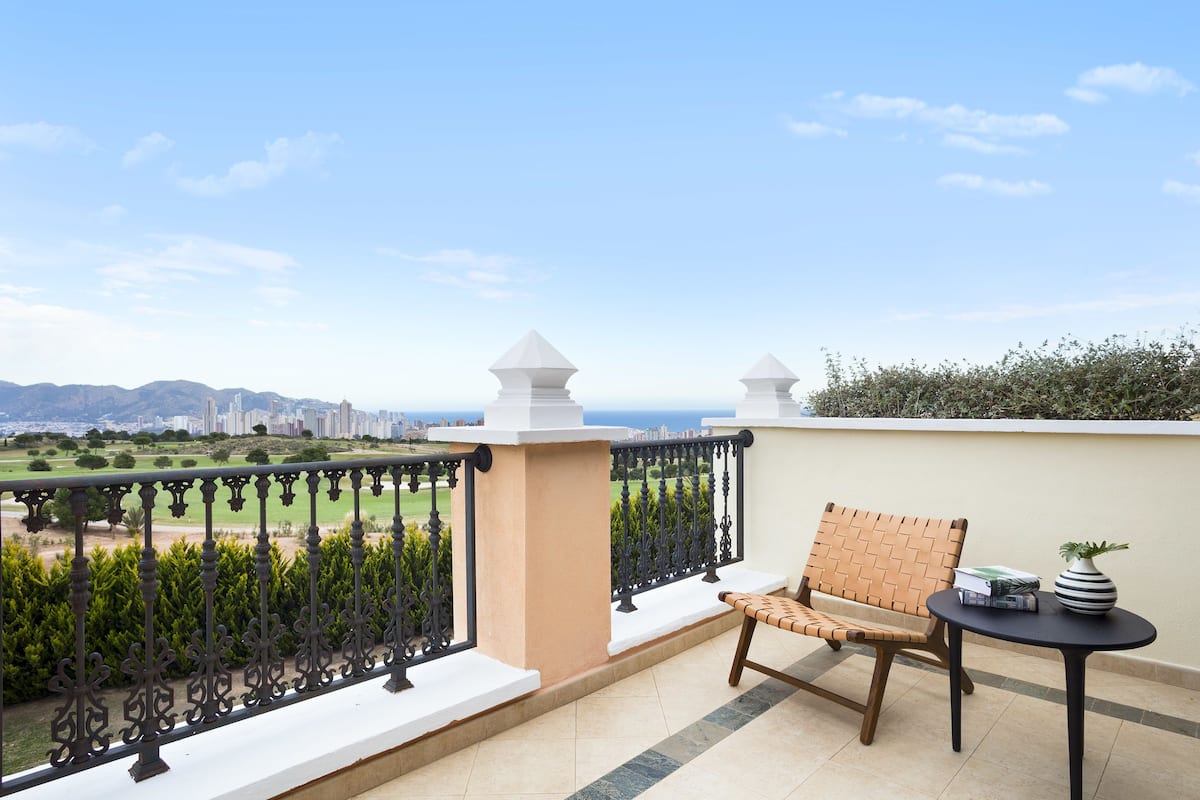 A balcony with a view of a cityscape, a wicker chair, and a small table with a vase.