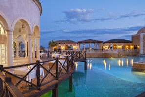 A poolside area with a wooden deck, tables with white tablecloths, and a building with arches and a tiled roof.