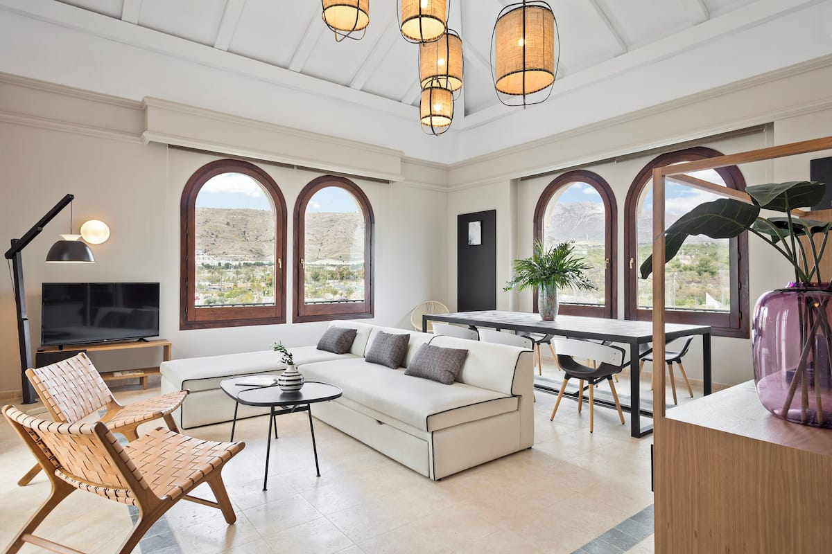 A modern living room with a sofa, chairs, a dining table, and large arched windows offering a view of the landscape.