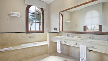A bathroom with a bathtub, two sinks, a large mirror, and a window with a view of palm trees.
