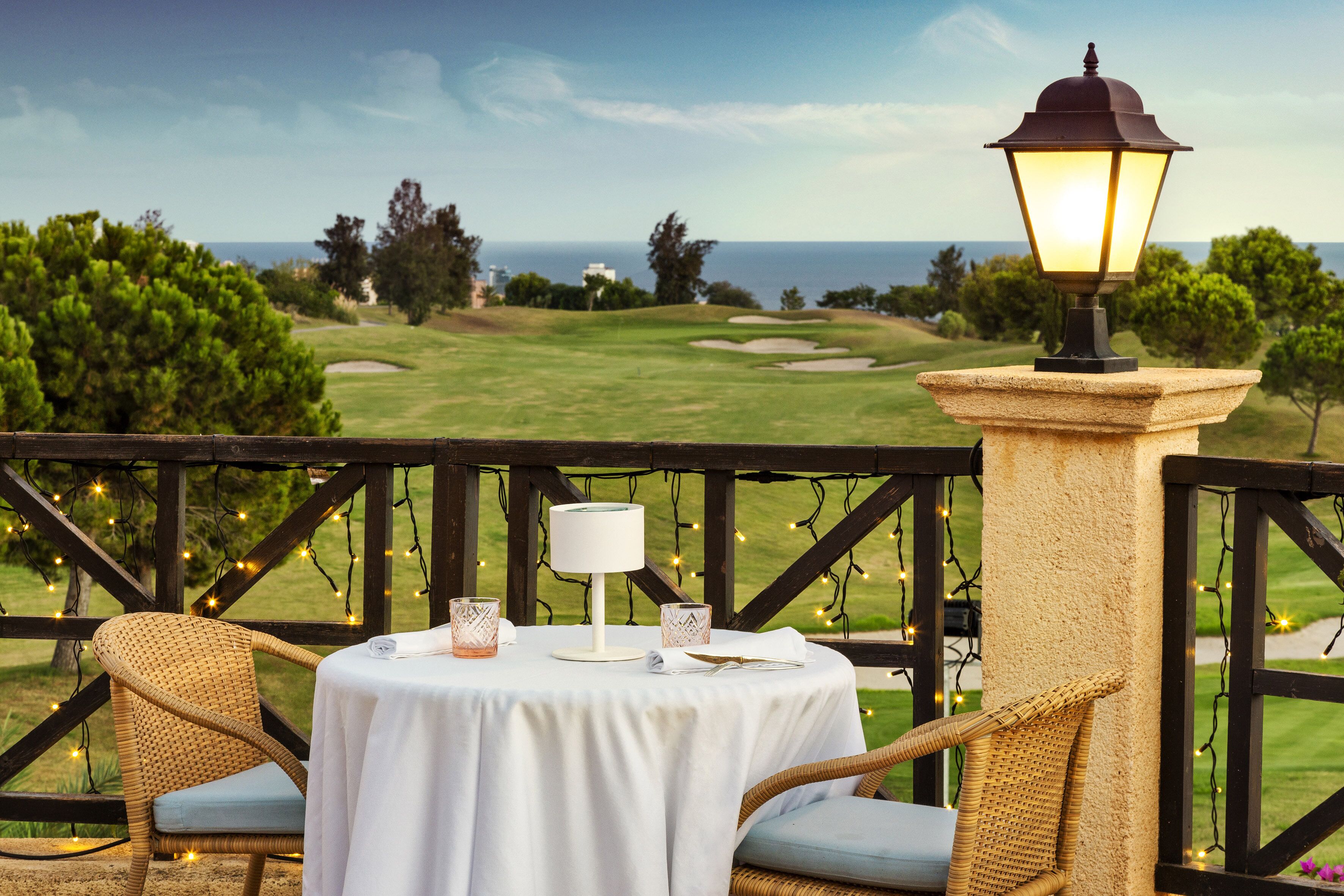 Una mesa redonda con mantel blanco, dos sillas de mimbre, una lámpara de mesa y luces decorativas en un balcón con vistas a un campo de golf.