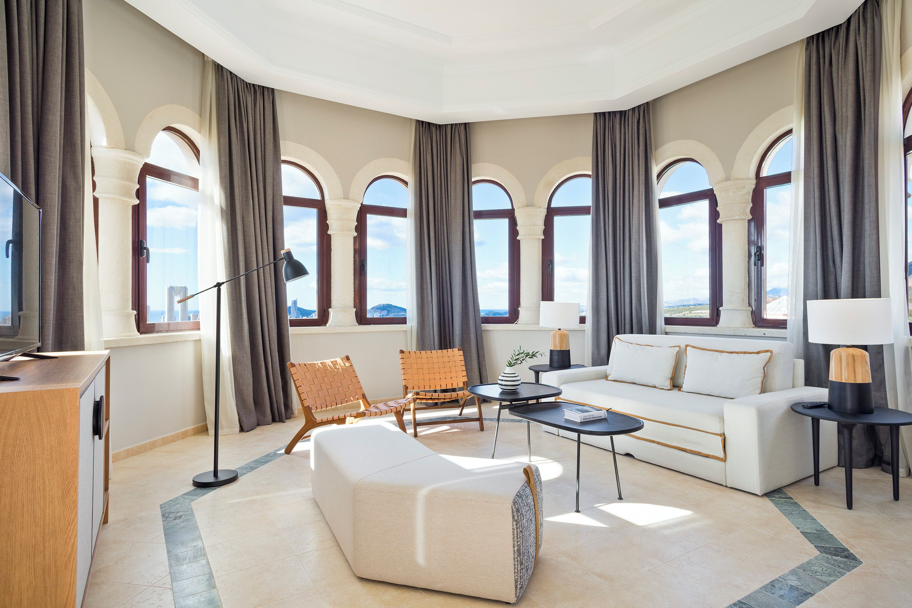 Amplia sala de estar con un sofá moderno, una mesa de centro y una zona de estar con un sillón. Amplias ventanas con cortinas ofrecen luz natural y vistas.