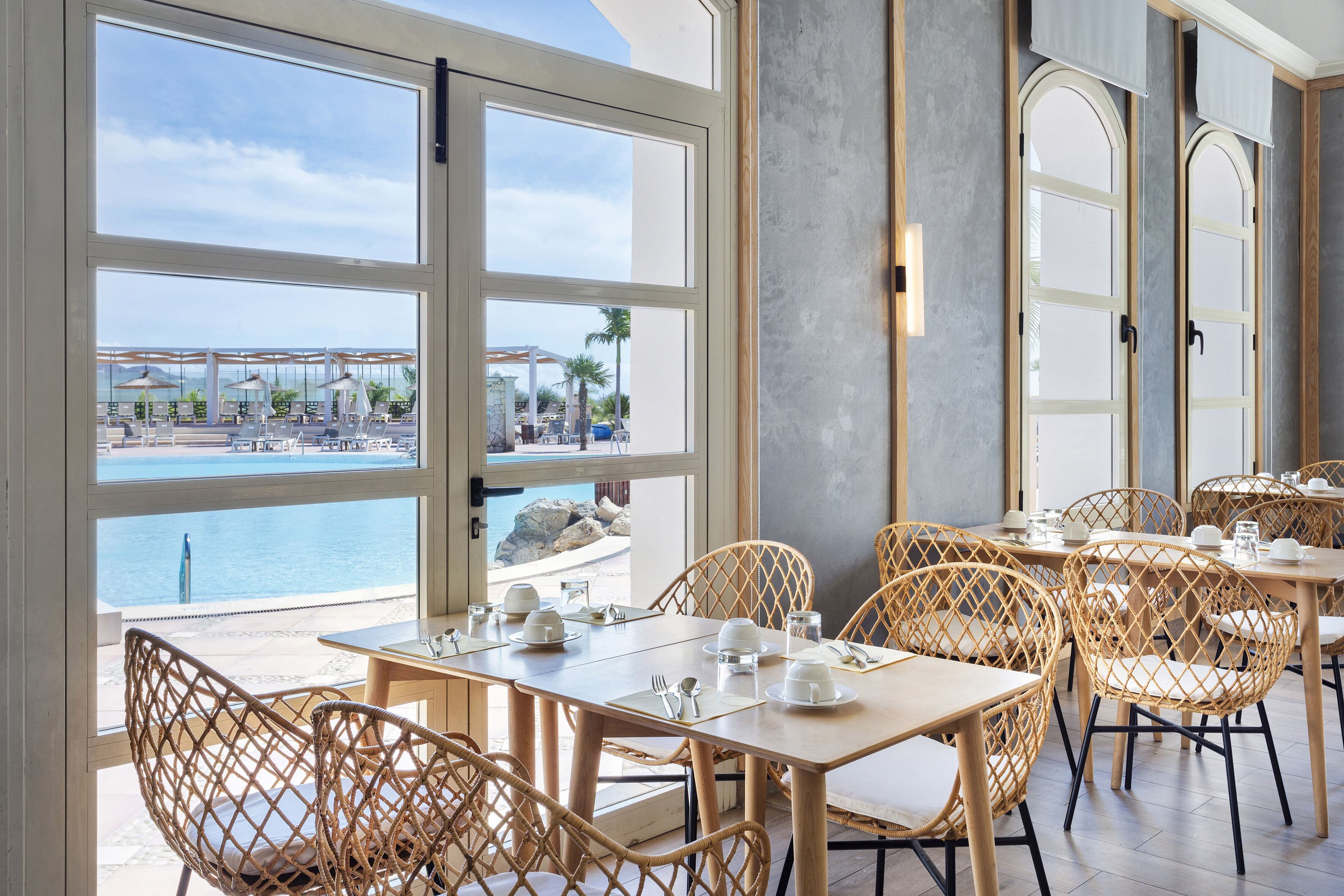 Un comedor con sillas y mesas de mimbre, ventanas amplias con vista a una piscina y una playa, y un diseño interior moderno.
