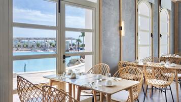 Un comedor con sillas y mesas de mimbre, ventanas amplias con vista a una piscina y una playa, y un diseño interior moderno.
