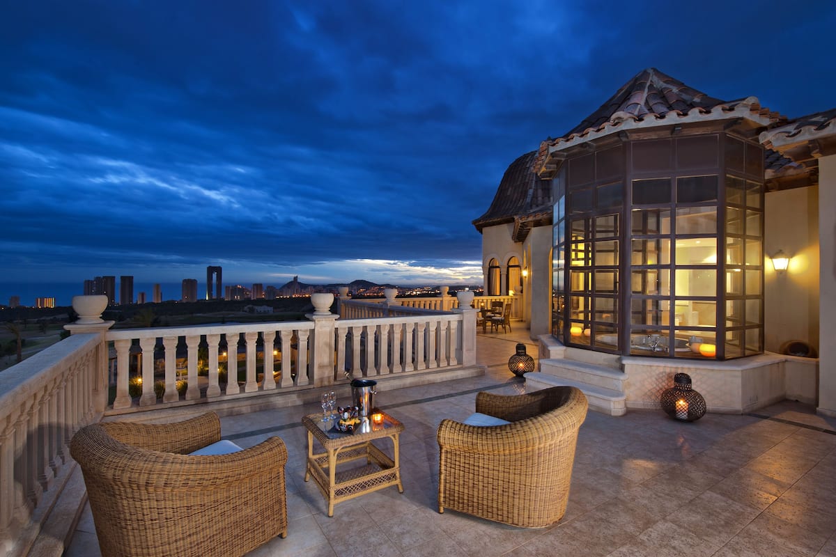 A rooftop terrace with wicker furniture, a glass-enclosed structure, and a city view at dusk.