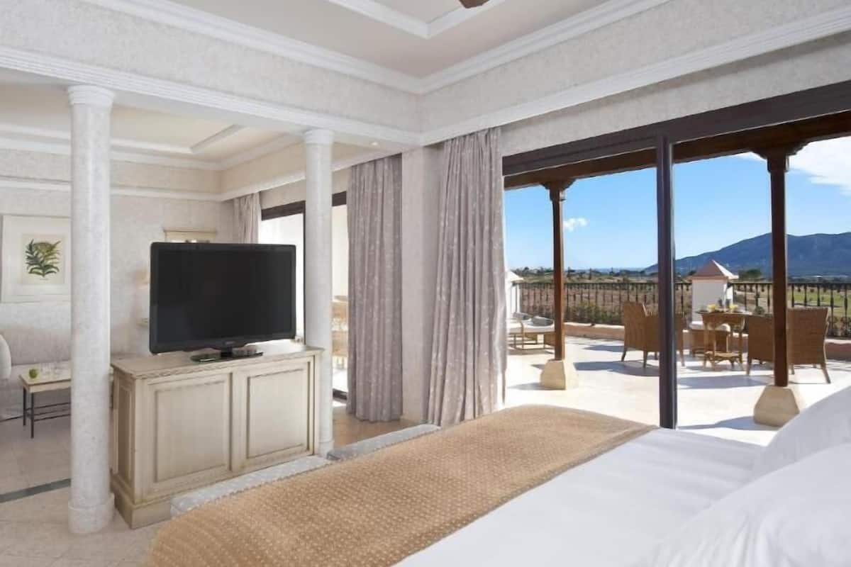 A hotel room with a bed, a TV on a cabinet, a balcony with outdoor furniture, and a view of the landscape.