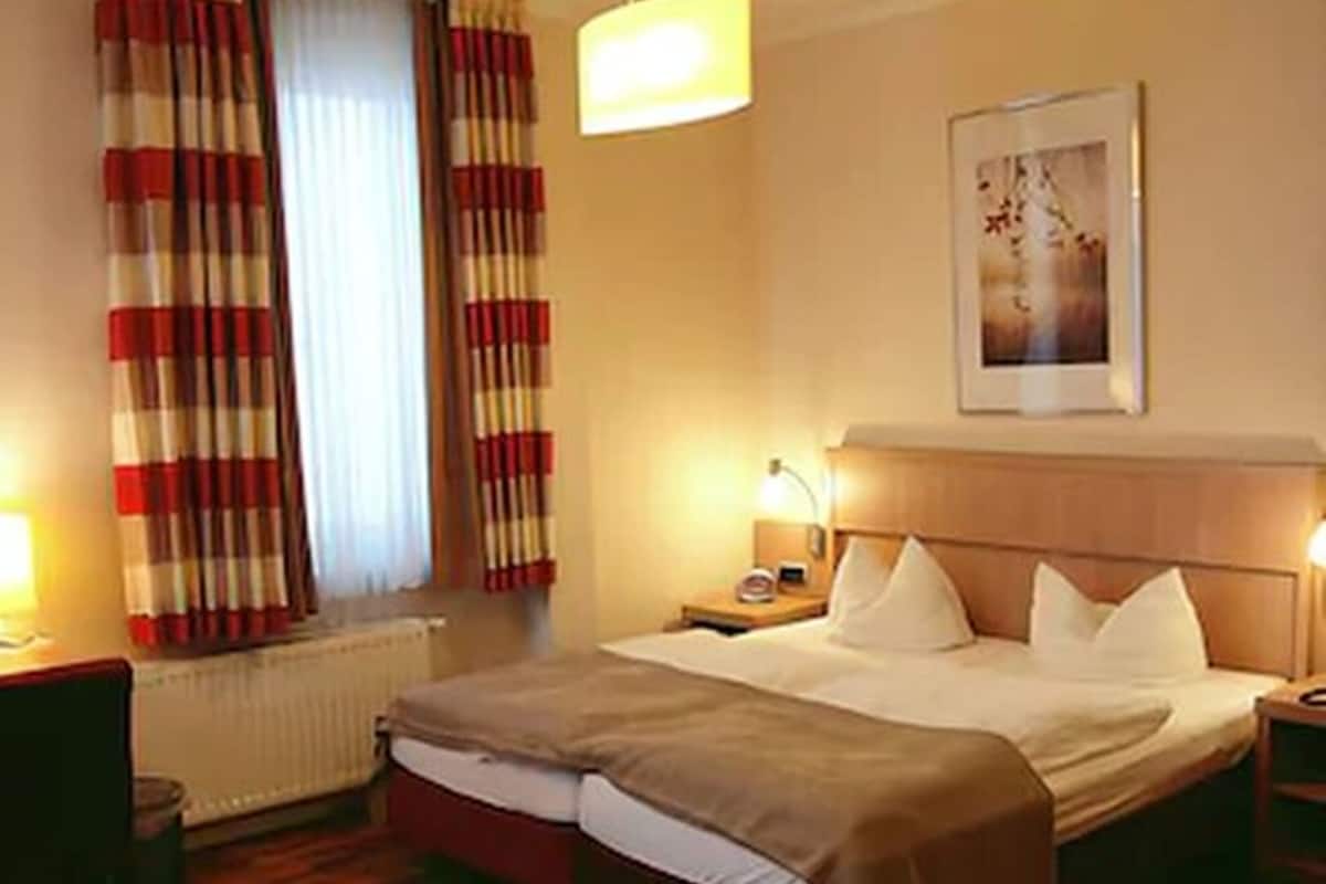 A hotel room with a bed, a nightstand, a lamp, a radiator, and curtains.