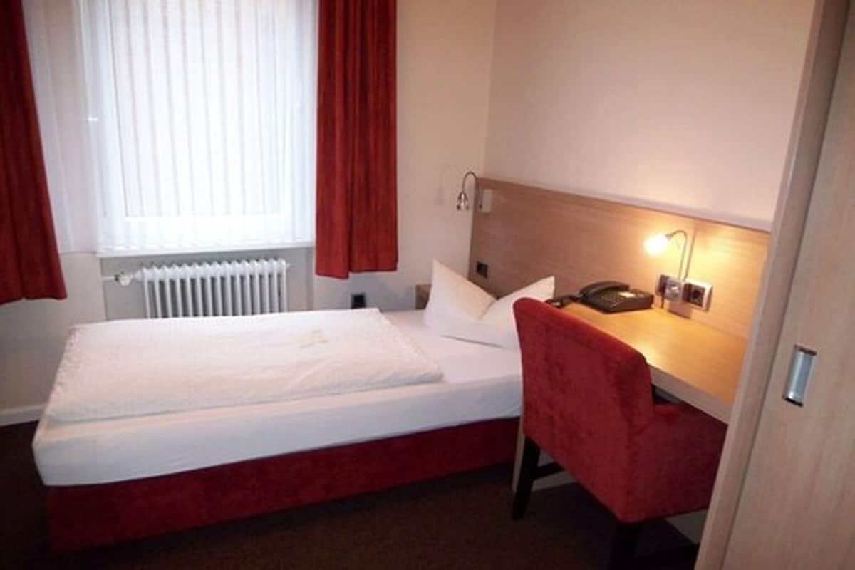 A hotel room with a single bed, a desk with a chair, a telephone, and a radiator.