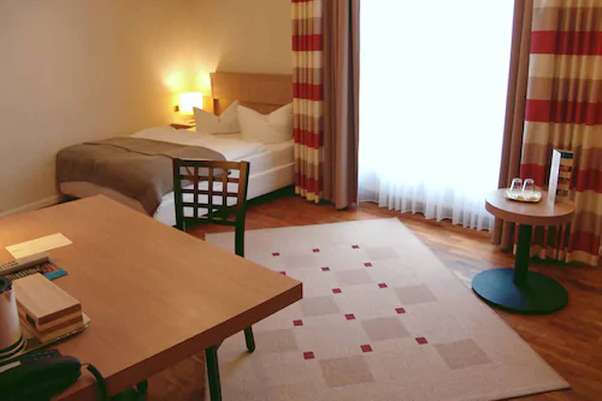 A hotel room with a bed, a desk, a chair, a small table, and a lamp.