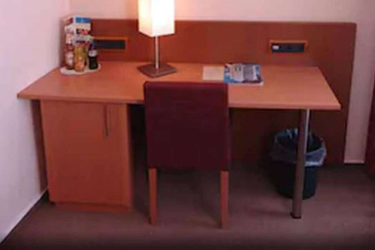 A small, simple room with a desk, chair, and a lamp. There is a trash bin and a framed picture on the wall.